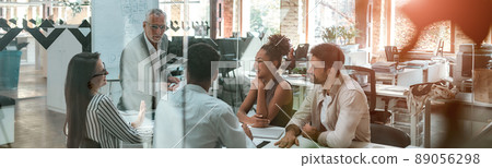 Business meeting. Mature businessman discussing something with his young colleagues while sitting together at the office table in the modern office 89056298