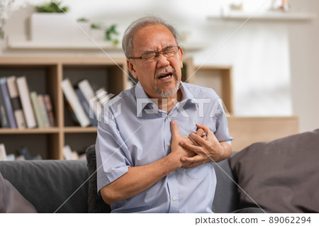 Elderly Asian male with from heart attack sitting at the couch in the living room. senior clutching his left chest from acute pain. Heart attack symptom, he has difficulty breathing. Elderly Asian male with from heart attack sitting at the couch in the living room. senior clutching his left chest from acute pain. Heart attack symptom, he has difficulty breathing. 89062294