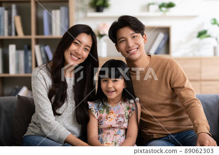 Happy Asian family sitting on couch smile with love and looking at camera spend time together.Father mother and little daughter relax on sofa in cozy living room.Good moment.Happy and warmth family. 89062303