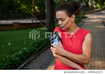Attractive stunning fit woman, athlete adjusting the mobile app in her mobile phone in the smartphone holder, standing in the city park, getting ready for morning jog Attractive stunning fit woman, athlete adjusting the mobile app in her mobile phone in the smartphone holder, standing in the city park, getting ready for morning jog 89062825