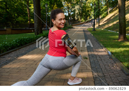 Rear view of an African woman doing lunges while exercising outdoor, smiles cutely with beautiful toothy smile looking at camera, enjoying her active and healthy lifestyle 89062829