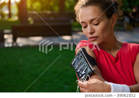 Close-up portrait of a beautiful woman, athlete adjusting the mobile app in her mobile phone in the smartphone holder, standing in the city park, getting ready for morning jog Close-up portrait of a beautiful woman, athlete adjusting the mobile app in her mobile phone in the smartphone holder, standing in the city park, getting ready for morning jog 89062852