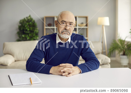 Portrait of serious senior man in glasses sitting at working desk and looking at camera Portrait of serious senior man in glasses sitting at working desk and looking at camera 89065098