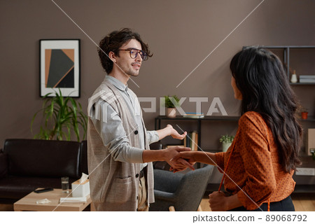 Horizontal medium shot of young psychologist greeting woman shaking hands and offering her to take seat Horizontal medium shot of young psychologist greeting woman shaking hands and offering her to take seat 89068792