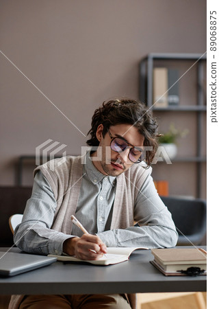 Vertical medium portrait of handsome young adult man wearing stylish casual outfit with eyeglasses sitting at desk writing something in notebook Vertical medium portrait of handsome young adult man wearing stylish casual outfit with eyeglasses sitting at desk writing something in notebook 89068875