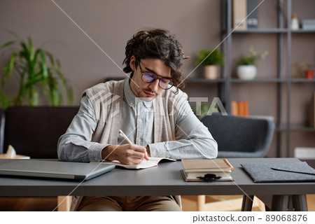 Horizontal medium portrait of young adult man wearing comfortable casual outfit and eyeglasses sitting at desk writing something in notebook Horizontal medium portrait of young adult man wearing comfortable casual outfit and eyeglasses sitting at desk writing something in notebook 89068876