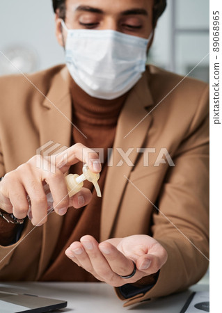 Young man in beige jacket sitting at table and using hand sanitizer in office 89068965