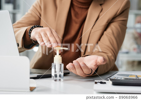 Close-up of unrecognizable man in stylish jacket sitting at desk and sterilizing hands with antiseptic 89068966