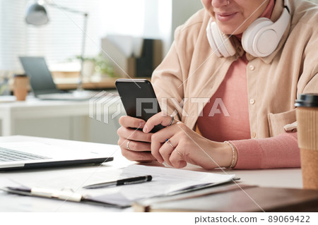Close-up of content modern young businesswoman in headphones around neck sitting at table in office and reading message on phone 89069422