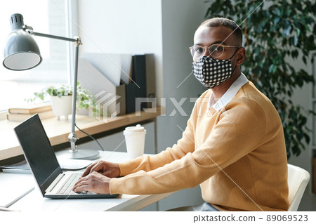 Portrait of serious young mixed race manager in checkered mask and eyeglasses sitting at table with laptop in office 89069523