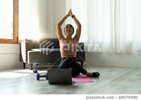 Calm relaxed young asian woman sitting in lotus position with hands up namaste gesture, closed eyes, breathing, resting, deeply meditating, practicing yoga on yoga mat at home 89070490