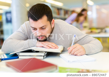 Portrait of upset adult man with stack of books on table in room of public library Portrait of upset adult man with stack of books on table in room of public library 89072447