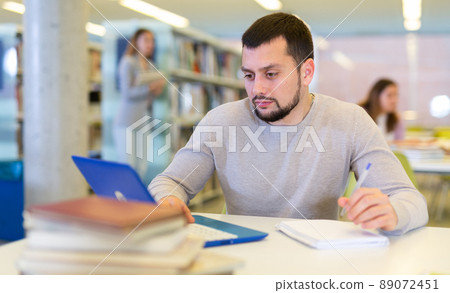 Middle aged man reading books and working on laptop in library Middle aged man reading books and working on laptop in library 89072451