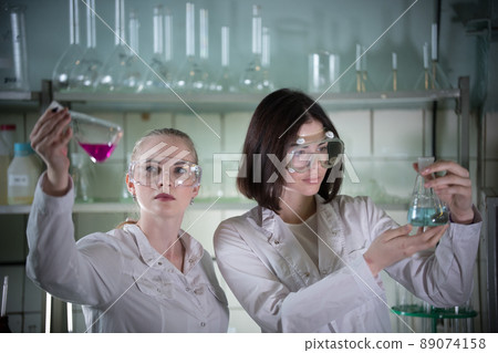 Chemical laboratory. Two young woman in work glasses holding different flasks with liquids in it. Looking at the flask Chemical laboratory. Two young woman in work glasses holding different flasks with liquids in it. Looking at the flask 89074158