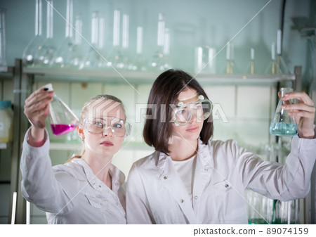 Chemical laboratory. Two young pretty woman holding different flasks with liquids in it. Looking at the flask 89074159