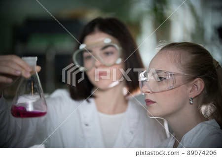 Chemical laboratory. Two young woman holding a flask with red liquid in it looking at it Chemical laboratory. Two young woman holding a flask with red liquid in it looking at it 89074206
