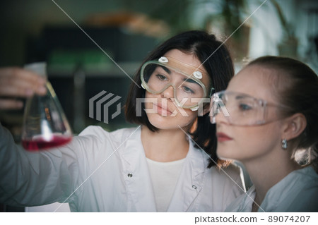 Chemical laboratory. Two young woman holding a flask with pink liquid in it looking at it Chemical laboratory. Two young woman holding a flask with pink liquid in it looking at it 89074207