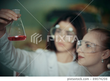 Chemical laboratory. Two young woman holding a flask with red liquid in it Chemical laboratory. Two young woman holding a flask with red liquid in it 89074208