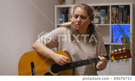 A girl with a serious expression on her face plays an acoustic guitar while sitting in a room. Behind her is a rack of board games and flowers. Musical composition. 89076732