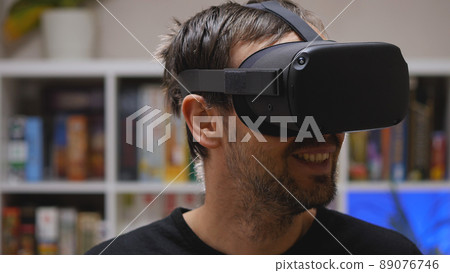 A bearded man wearing VR glasses smiles at the virtual world he sees. Close-up. In the background is a rack of games. VR helmet for entertainment, education, and gaming. A bearded man wearing VR glasses smiles at the virtual world he sees. Close-up. In the background is a rack of games. VR helmet for entertainment, education, and gaming. 89076746
