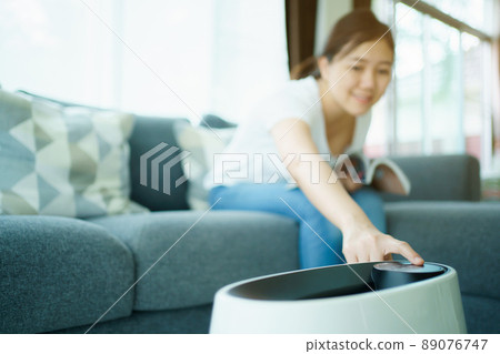 Happy Asian young woman turning on high efficiency air purifier while staying and reading a book in the living room. Woman using air purifier in her house. 89076747