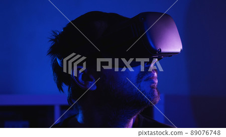 Close-up of a bearded man wearing VR glasses in a dark room with blue light. There is a room flower in the front captivity. VR helmet for entertainment, learning and gaming. Close-up of a bearded man wearing VR glasses in a dark room with blue light. There is a room flower in the front captivity. VR helmet for entertainment, learning and gaming. 89076748