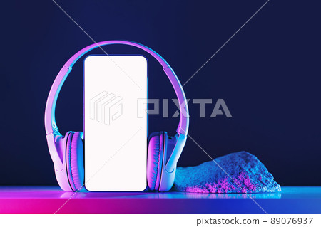 Smartphone mockup display with grey wireless headphones on neon background Smartphone mockup display with grey wireless headphones on neon background 89076937