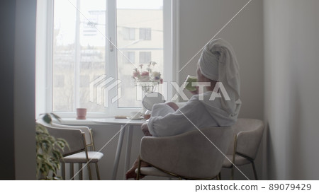 A young woman in a bathrobe and with a towel on her head, made herself a smoothie. The girl is sitting in the kitchen and has a smoothie for breakfast. Lifestyle A young woman in a bathrobe and with a towel on her head, made herself a smoothie. The girl is sitting in the kitchen and has a smoothie for breakfast. Lifestyle 89079429