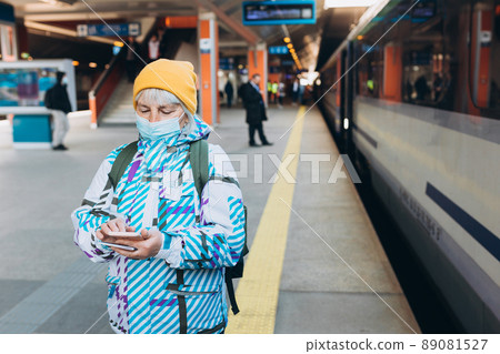 Old woman in yellow hat waiting on station platform with backpack on background train using smart phone. Railroad transport concept, Traveler 89081527
