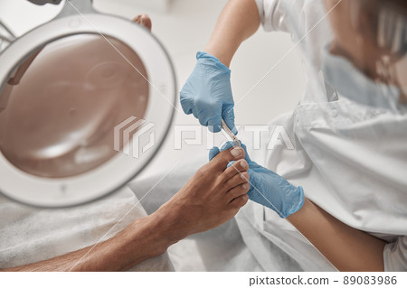 Skilled podiatrist in gloves cuts toenail of client with clipper in salon Skilled podiatrist in gloves cuts toenail of client with clipper in salon 89083986