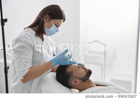 Woman in mask does filler injection for eyebrow lifting to middle aged man in salon Woman in mask does filler injection for eyebrow lifting to middle aged man in salon 89083987