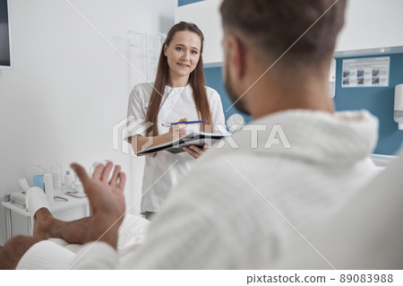 Man client in bathrobe talks to positive woman holding clipboard in beauty salon Man client in bathrobe talks to positive woman holding clipboard in beauty salon 89083988