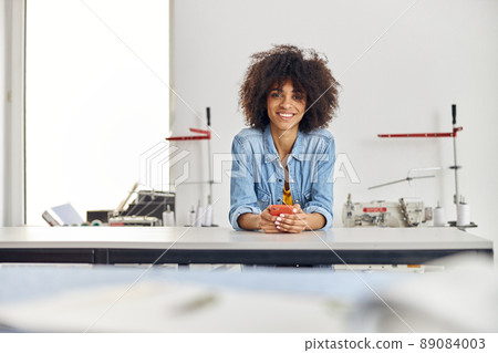 Portrait of smiling African-American woman with mobile phone leaning onto cutting table Portrait of smiling African-American woman with mobile phone leaning onto cutting table 89084003