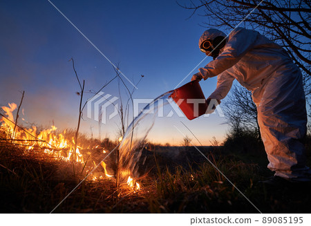 Research scientist fighting fire in field with blue night sky on background. Man in protective radiation suit and gas mask holding bucket and pouring water on burning grass with smoke. Research scientist fighting fire in field with blue night sky on background. Man in protective radiation suit and gas mask holding bucket and pouring water on burning grass with smoke. 89085195