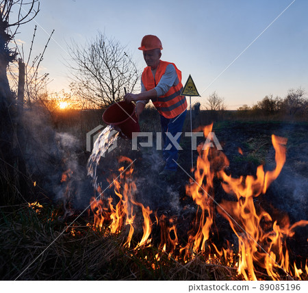 Fireman ecologist fighting fire in field with evening sky on background. Man holding bucket and pouring water on burning dry grass near yellow triangle with skull and crossbones warning sign. Fireman ecologist fighting fire in field with evening sky on background. Man holding bucket and pouring water on burning dry grass near yellow triangle with skull and crossbones warning sign. 89085196