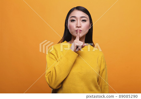 Beautiful mysterious woman wearing vibrant yellow sweater making silence gesture on orange background. Secretive person touching lips with forefinger indicating silence and secrecy. 89085290
