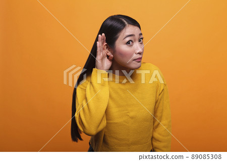 Curious brunette asian woman eavesdropping interesting gossip and secret rumours about her. Intrigued serious person with hand at ear overhearing people gossiping with interest. 89085308