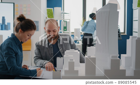 Team of coworkers analyzing building model and blueprints plan on table to design construction layout. Architectural colleagues doing teamwork, designing project development together. Team of coworkers analyzing building model and blueprints plan on table to design construction layout. Architectural colleagues doing teamwork, designing project development together. 89085545