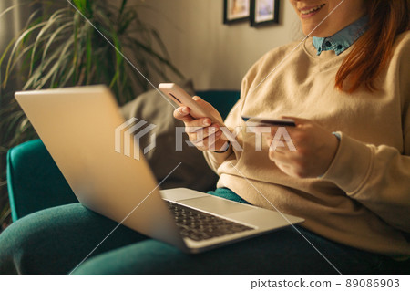 woman holding credit card and using laptop and smartphone sits on sofa at home. Business credit banking and online shopping service woman holding credit card and using laptop and smartphone sits on sofa at home. Business credit banking and online shopping service 89086903