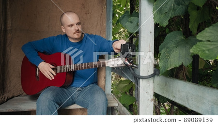 Man with an acoustic guitar is sitting on porch of farmhouse, tuning instrument using an app on his phone. Concept of creativity, hobbies, lifestyle and recreation. Old, retro decor, burlap background Man with an acoustic guitar is sitting on porch of farmhouse, tuning instrument using an app on his phone. Concept of creativity, hobbies, lifestyle and recreation. Old, retro decor, burlap background 89089139
