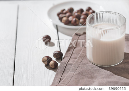 dairy substitution, healthy plant milk in a glass. hazelnut drink on a white wooden table next to nuts on a plate.. 89090633