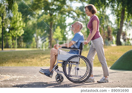 A woman carrying her husband in a wheel chair in a park A woman carrying her husband in a wheel chair in a park 89091690