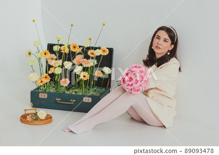 Beautiful brunette girl in a white jacket and white tights in a light interior holding a rose bouquet. Suitcase with flowers on the background Beautiful brunette girl in a white jacket and white tights in a light interior holding a rose bouquet. Suitcase with flowers on the background 89093478