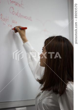 Profile view of a cute young brunette english teacher in white blouse writing on a board and giving a class. 89093570
