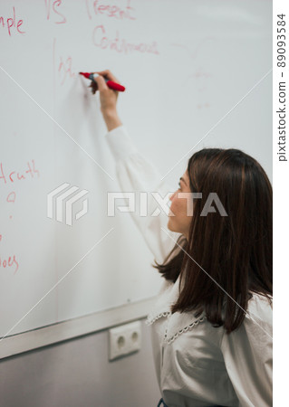 Profile view of a cute young brunette english teacher in white blouse writing on a board and giving a class. 89093584