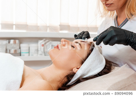 Professional cosmetology. Cosmetologist in latex gloves cleans the client's face using an ultrasound machine. Side view. Concept of beauty procedures Professional cosmetology. Cosmetologist in latex gloves cleans the client's face using an ultrasound machine. Side view. Concept of beauty procedures 89094096