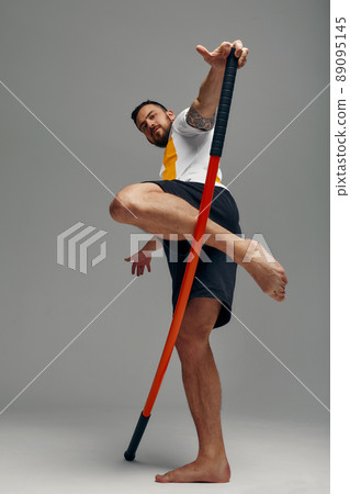 A fighter in white t-shirt and black shorts, stands in a fighting artificially pose and holds a fight stick on studio background. 89095145