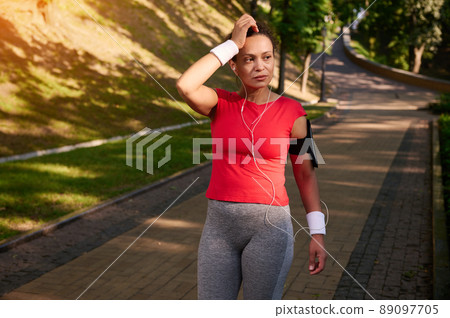 Exhausted tired African American woman, female athlete jogger runner walking on the city park footpath, relaxing after morning run. Exhausted tired African American woman, female athlete jogger runner walking on the city park footpath, relaxing after morning run. 89097705