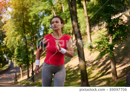 Attractive strength muscular multi ethnic woman in tight sportswear with earphones and smartphone holder running on the footpath in the city park 89097706