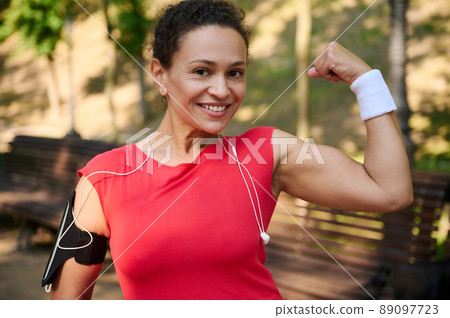 Active middle-aged sportswoman smiling beautiful toothy smile and showing her toned biceps to camera during workout. Push beyond your limits. 89097723